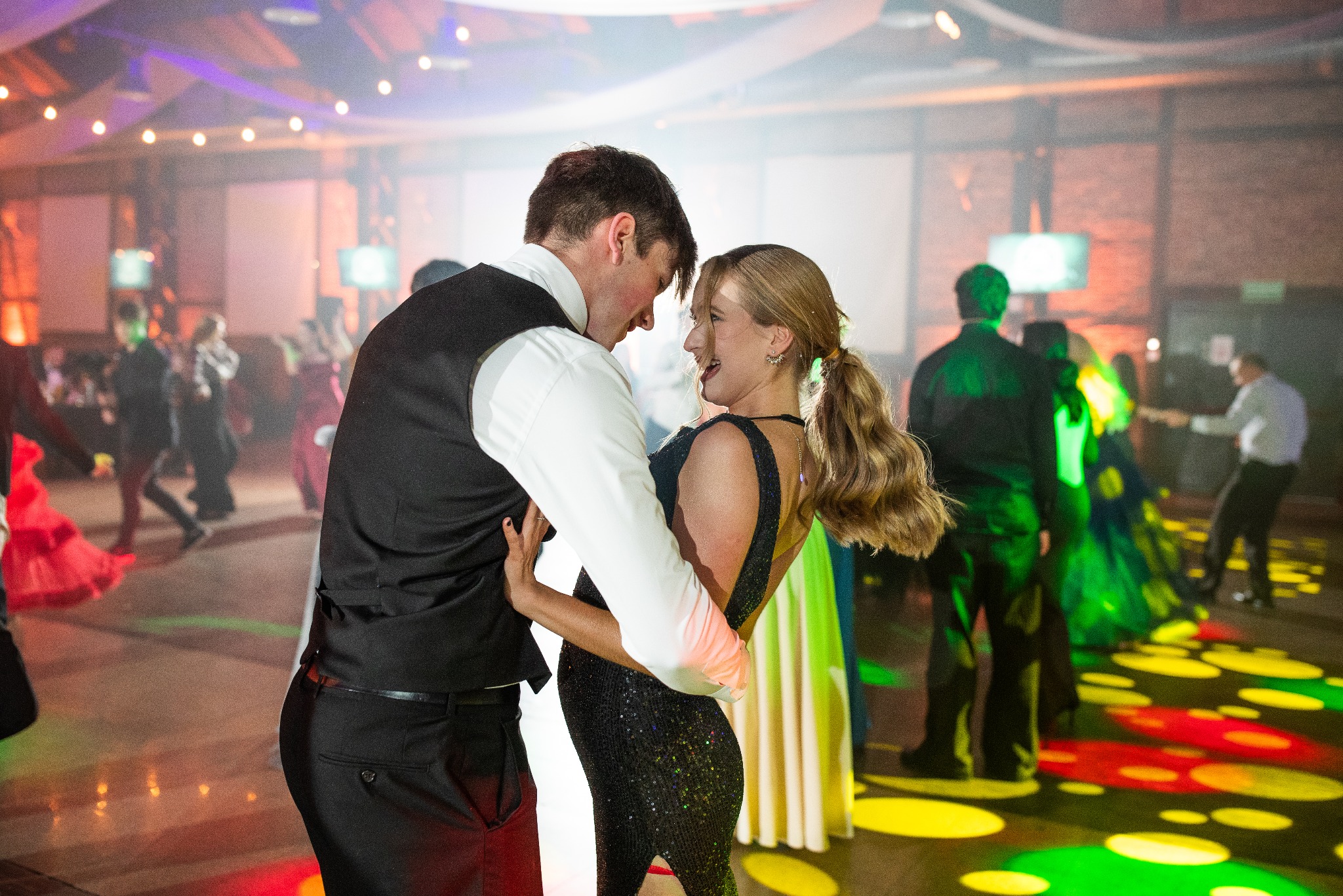Para tańczy na studniówce, ona w cekinowej sukience, on w kamizelce. Kolorowe światła na parkiecie, w tle inni tańczący.