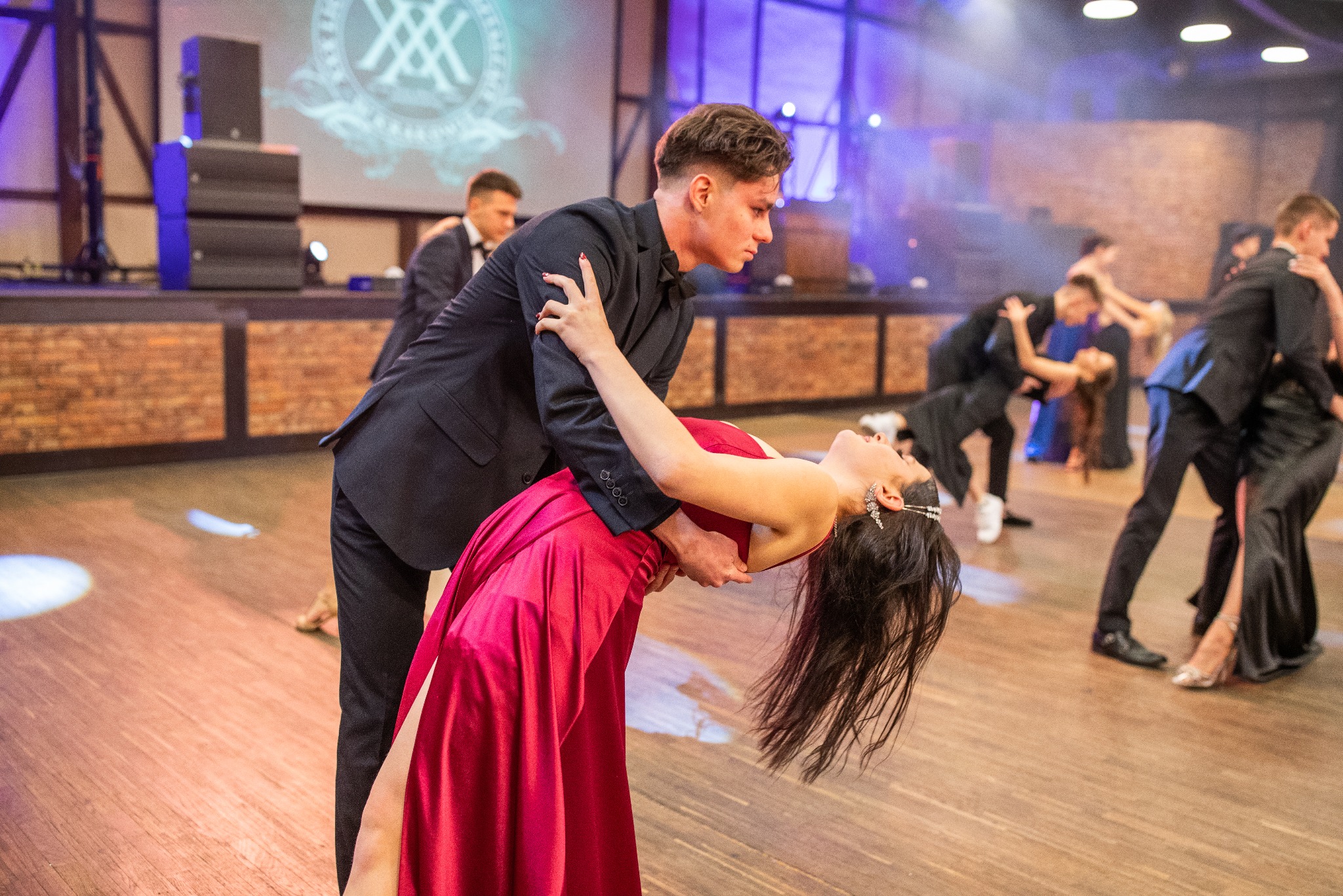 Para tańczy na parkiecie podczas studniówki. Dziewczyna w czerwonej sukni odchylona do tyłu, chłopak w czarnym garniturze ją podtrzymuje. W tle inne pary i oświetlenie sceniczne.