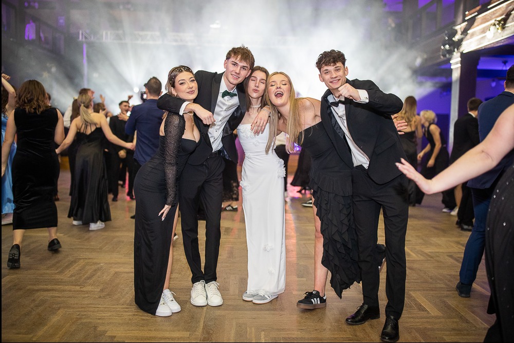 Radosna fotografia grupowa z balu studniówkowego w Krakowie. Młodzież w eleganckich strojach pozuje na parkiecie, w tle tańczący tłum i światła sceniczne.