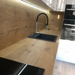 Nowoczesna kuchnia z drewnianym blatem i panelem ściennym imitującym drewno, czarny zlew i bateria, białe szafki z chromowanymi uchwytami oraz piekarnik w zabudowie.