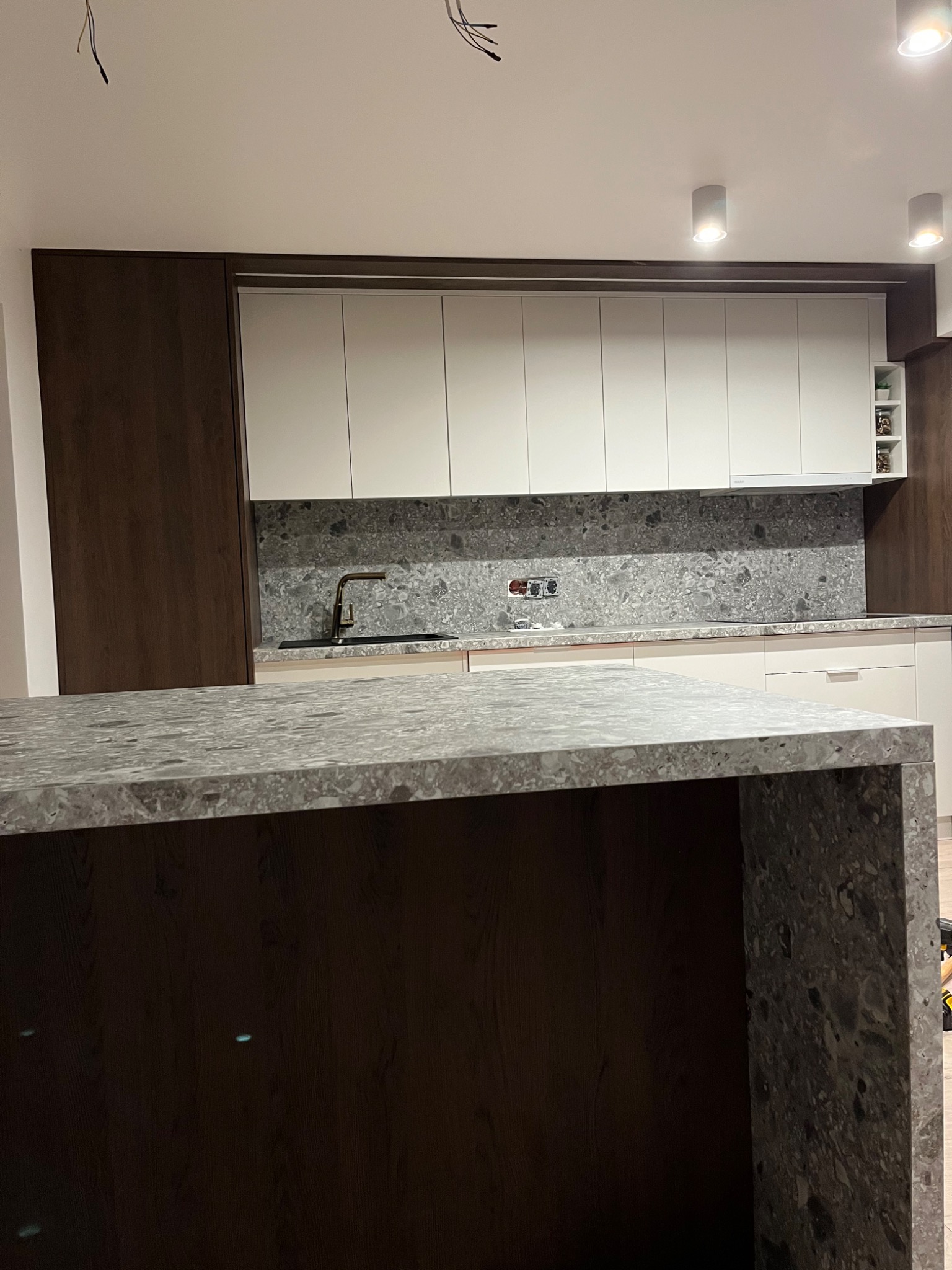 Nowoczesna kuchnia z wyspą: fronty szafek w kolorze złamanej bieli, ciemnobrązowe panele boczne i blat wyspy z szarego kamienia z wyraźnym wzorem, czarna bateria kuchenna, szary kamienny backsplash.