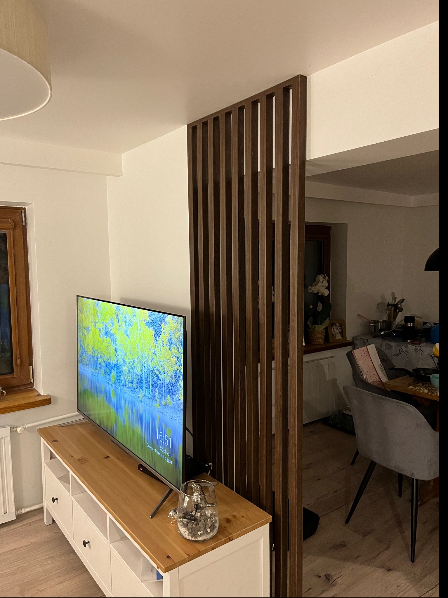 Drewniana ścianka działowa z pionowych listew w kolorze orzecha, oddzielająca salon od jadalni, widoczna obok telewizora na komodzie.