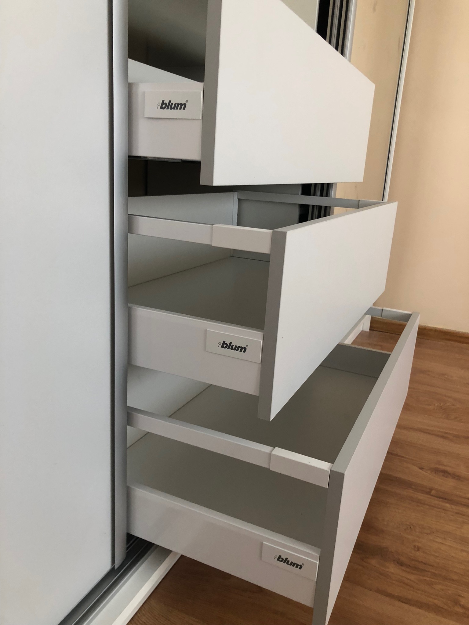 Wysuwane, białe szuflady z systemem Blum w szafie wnękowej z przesuwnymi drzwiami, widok z boku, parkiet na podłodze.