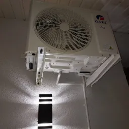 Biała jednostka zewnętrzna klimatyzatora marki Gree zamontowana na ścianie, widoczne rury odprowadzające skropliny i oświetlenie ścienne.