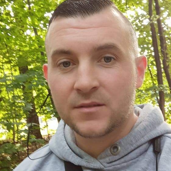 Portret mężczyzny w szarej bluzie z kapturem na tle zieleni drzew