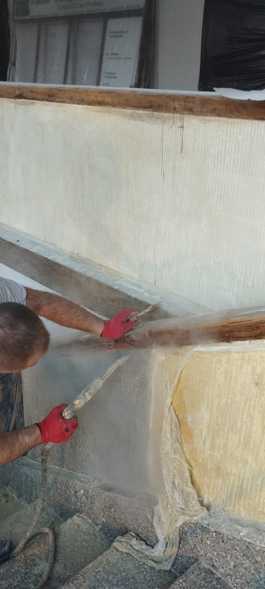 Pracownik w czerwonych rękawicach czyści ścianę budynku za pomocą piaskarki, zabezpieczone elementy drewniane i folia ochronna na ścianie.