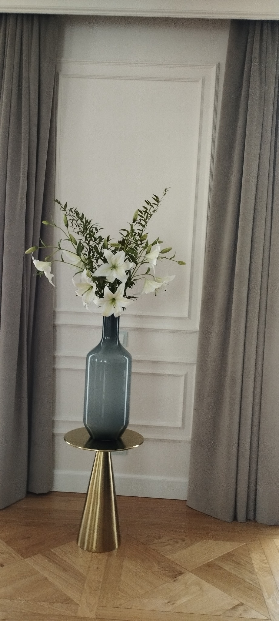 Elegancki bukiet białych lilii w szklanym, szarym wazonie na złotej, stożkowej podstawie, ustawiony na parkiecie w jodełkę, na tle białej ściany z dekoracyjnymi listwami i szarych zasłon.