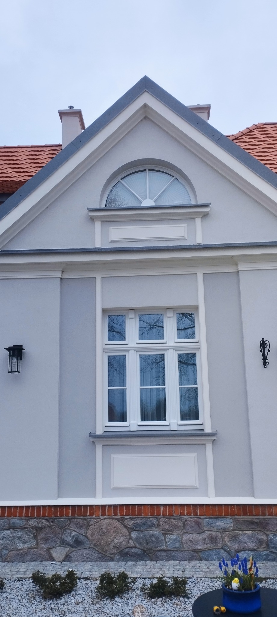 Elewacja budynku z białymi oknami, półokrągłym oknem na szczycie i dekoracyjnymi lampami ściennymi, wykończona kamienną podmurówką z ceglanym pasem.