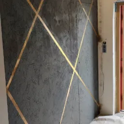 Szara ściana z dekoracyjnym tynkiem strukturalnym i złotymi, ukośnymi pasami, widoczny fragment podłogi zabezpieczonej folią malarską, gniazdko elektryczne i fragment futryny drzwi.