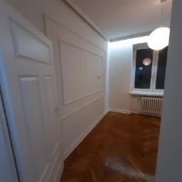 Pokój po renowacji z parkietem w jodełkę, białe ściany z dekoracyjnymi listwami, białe drzwi i okno z grzejnikiem, oświetlony lampą sufitową.