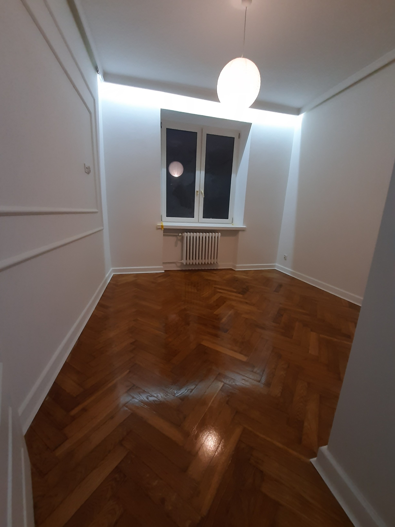 Pusty pokój po odświeżeniu: parkiet w jodełkę, białe ściany z dekoracyjnymi listwami, grzejnik pod oknem, okrągła lampa sufitowa i oświetlenie LED ukryte nad listwami.