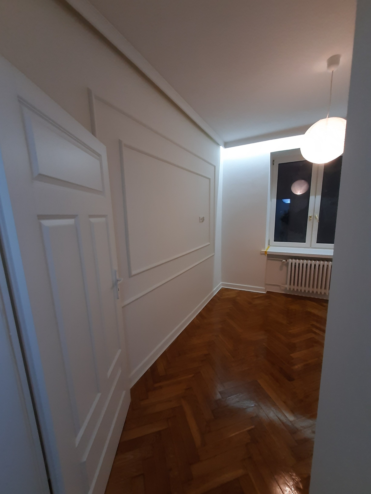 Pokój po renowacji z parkietem w jodełkę, białe ściany z dekoracyjnymi listwami, białe drzwi i okno z grzejnikiem, oświetlony lampą sufitową.