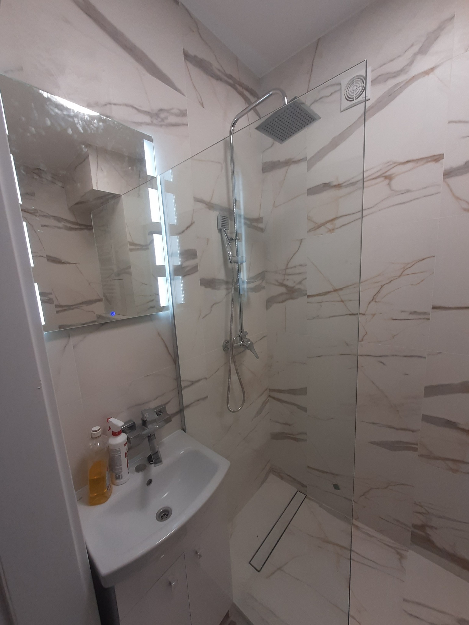 Nowoczesna łazienka z kabiną prysznicową typu walk-in, umywalką nablatową i lustrem z podświetleniem LED, wykończona płytkami imitującymi marmur z brązowymi żyłami.