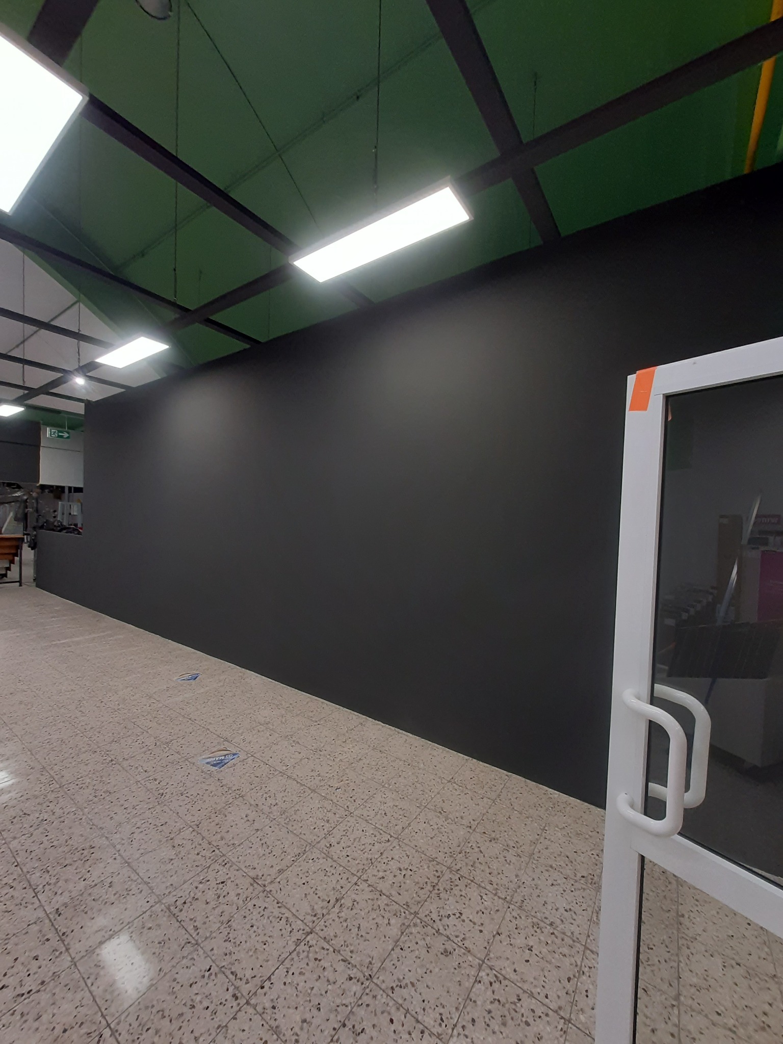 Świeżo pomalowana, matowa, czarna ściana w pomieszczeniu z terakotową podłogą i białymi drzwiami z taśmą zabezpieczającą na framudze.