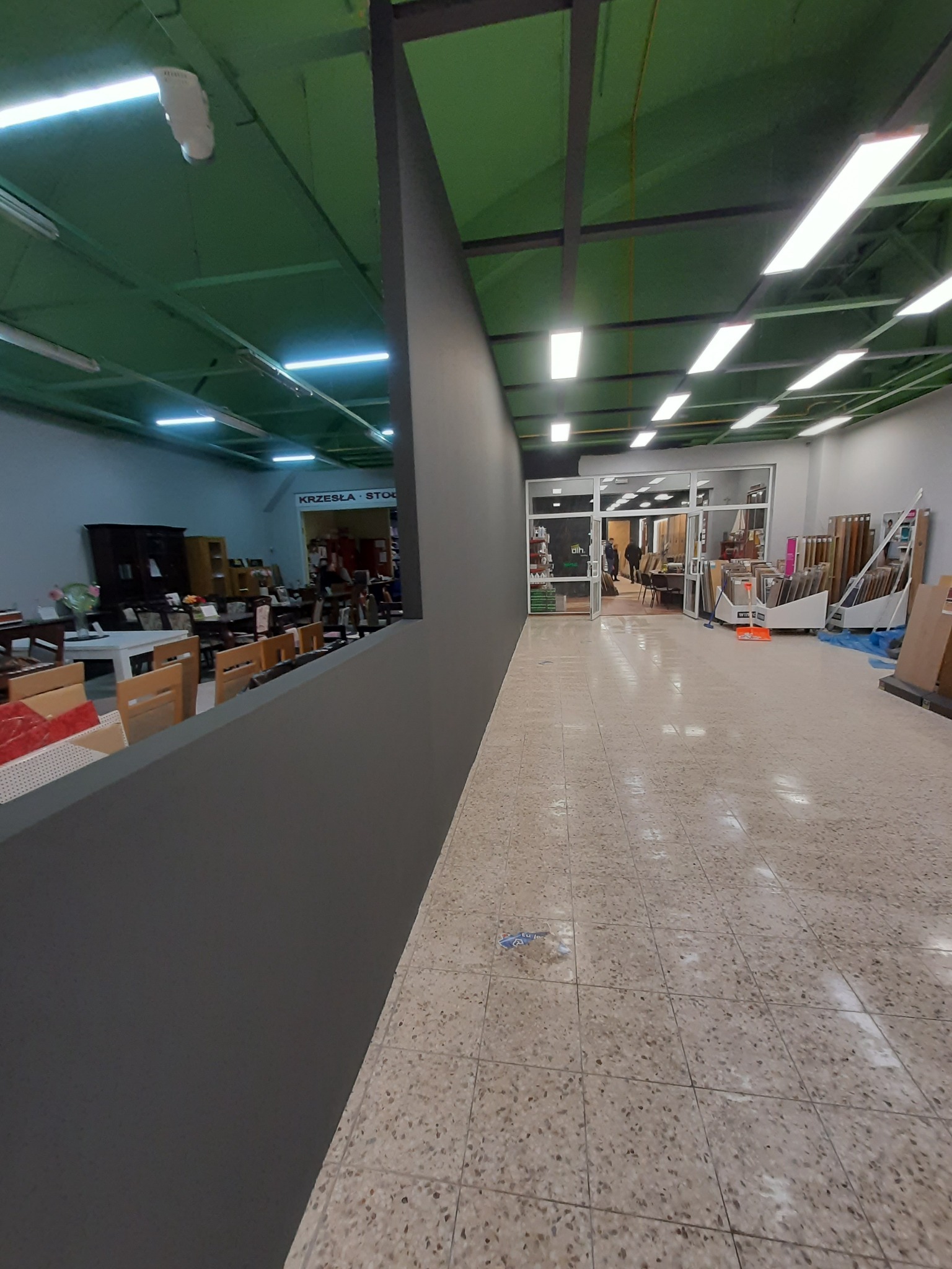 Wnętrze sklepu meblowego z widocznymi krzesłami i stołami za szarą ścianą działową, oświetlone jarzeniówkami pod zielonym sufitem.