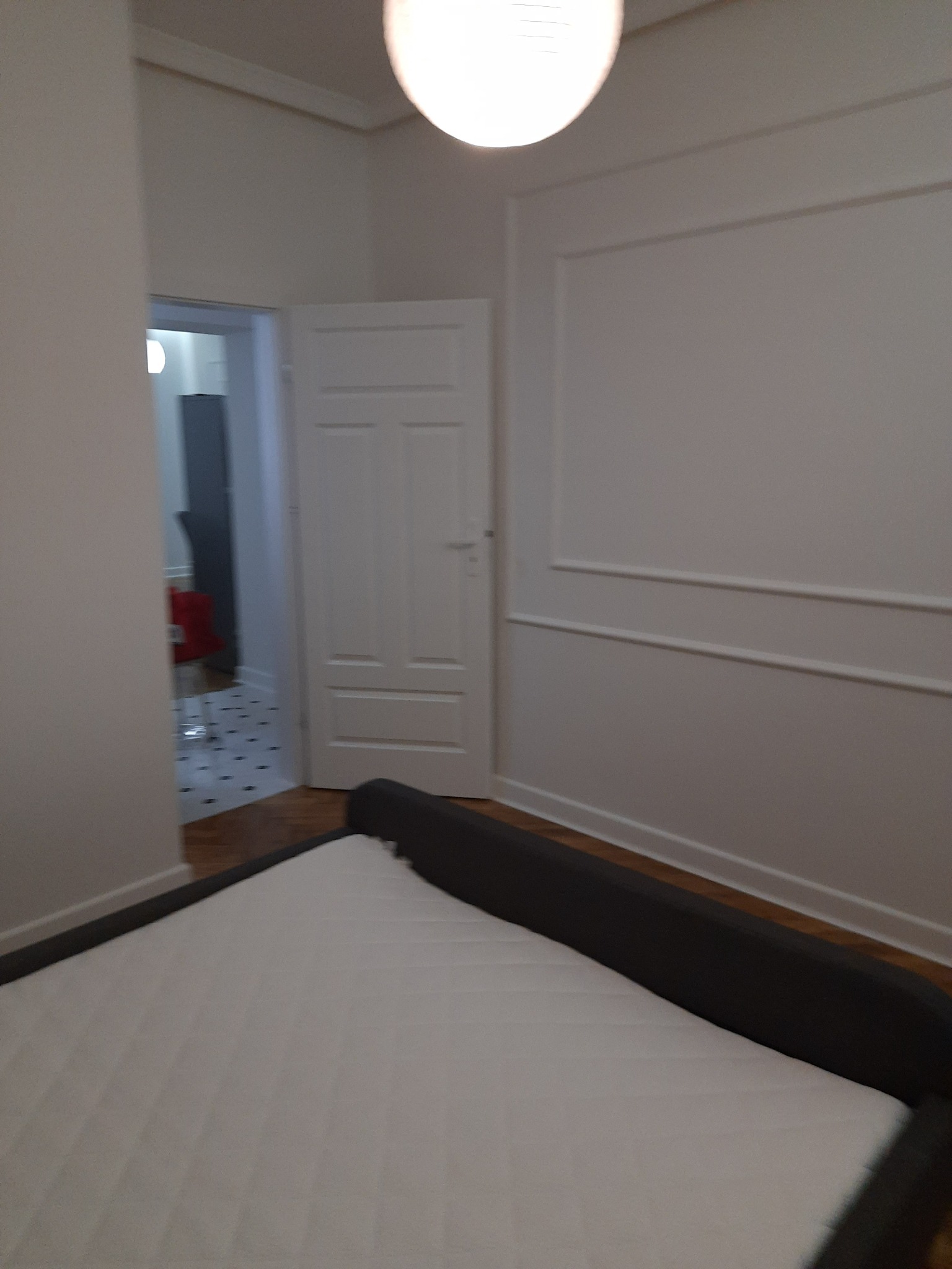 Pomalowane na biało ściany sypialni z dekoracyjnymi listwami, widoczny fragment łóżka z szarym wezgłowiem i otwarte drzwi do pokoju z czerwonym krzesłem i wzorzystą podłogą.