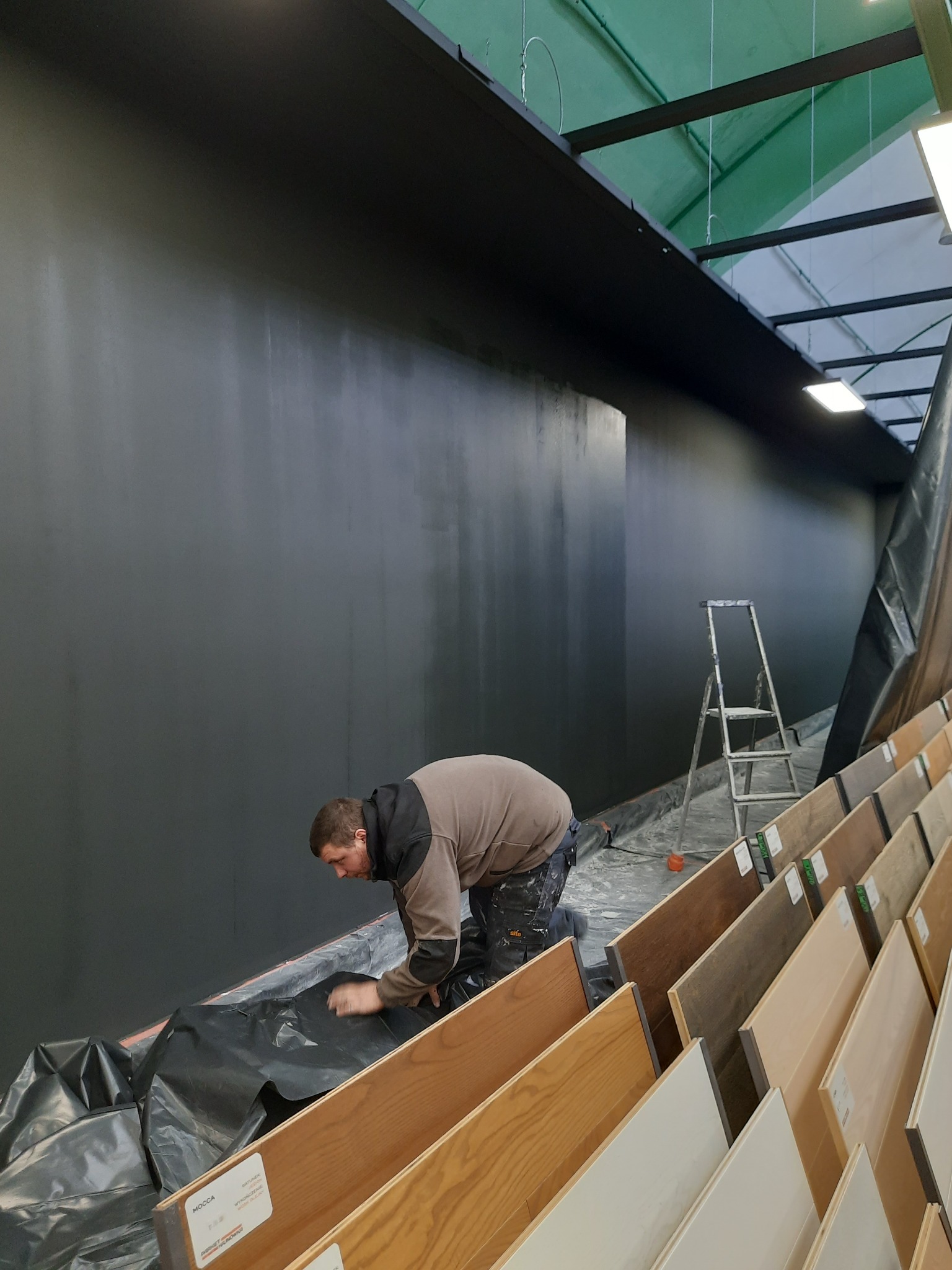 Mężczyzna w trakcie malowania ściany na ciemny kolor w sklepie z panelami podłogowymi, widoczna drabina i zabezpieczona folią podłoga.