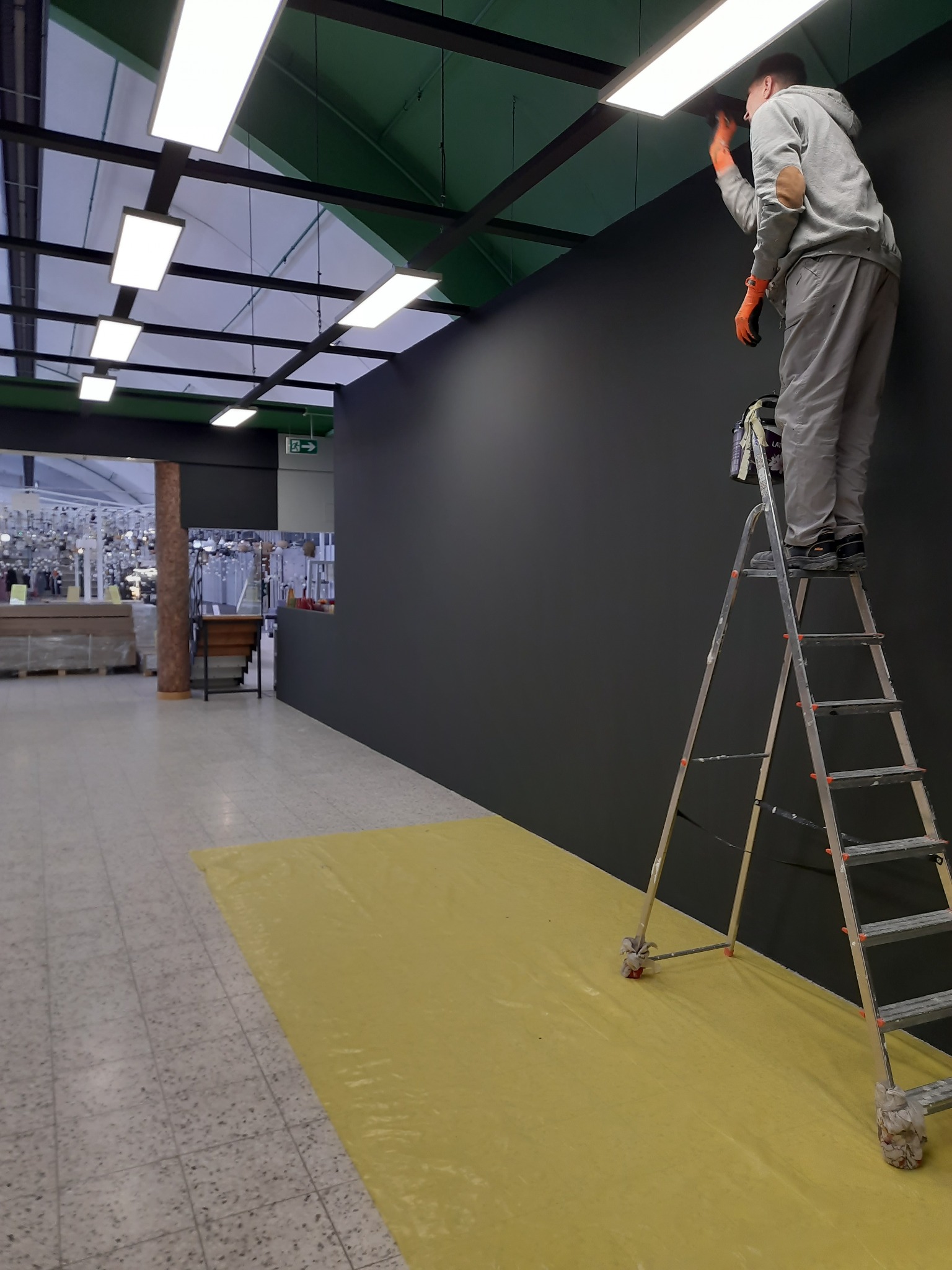 Mężczyzna w pomarańczowych rękawicach maluje ciemną ścianę w pomieszczeniu handlowym, stojąc na aluminiowej drabinie z zabezpieczonymi nogami, podłoga zabezpieczona żółtą folią, widoczne...