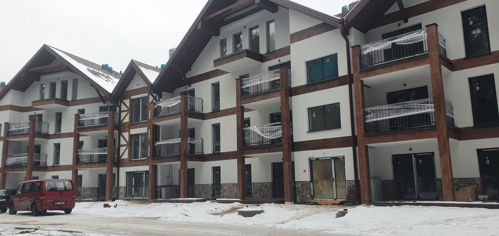 Nowe, trzypiętrowe apartamenty z balkonami zabezpieczonymi folią, drewnianymi elementami konstrukcyjnymi i kamienną okładziną na parterze, częściowo zasypane śniegiem, z czerwonym vanem...