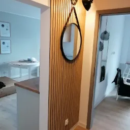 Drewniana ścianka działowa z pionowych listew, ozdobiona okrągłym lustrem na skórzanym pasku i czarną lampą, widok na wnętrze mieszkania z białymi ścianami i drewnianą podłogą.