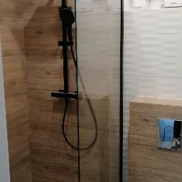 Nowoczesna kabina prysznicowa typu walk-in z czarną armaturą, drewnopodobne płytki na ścianach, odpływ liniowy w podłodze, widoczny fragment toalety i okna.