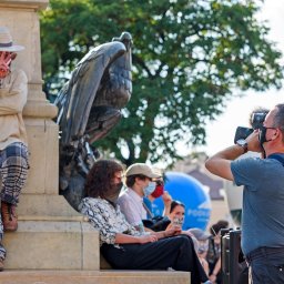 Transmisje na żywo - VisionRed.pl - live streaming online - Operator kamery z profesjonalnym sprzętem filmuje osoby siedzące obok pomnika orła. Jedna osoba oparta o pomnik w słomkowym kapeluszu pokazuje znak zwycięstwa.