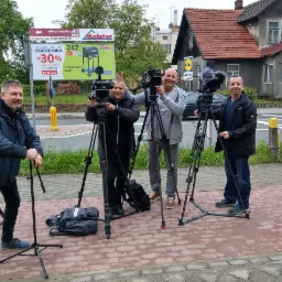 Czterech mężczyzn z profesjonalnymi kamerami na statywach, prawdopodobnie podczas kręcenia materiału w plenerze, billboardy i budynek w tle.