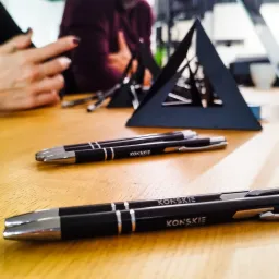 Dwa czarne długopisy z srebrnymi elementami i napisem 'KONSKIE' leżą na jasnym drewnianym biurku obok czarnej, trójkątnej podstawki na wizytówki. W tle widoczne dłonie i fragmenty ubrań osób.