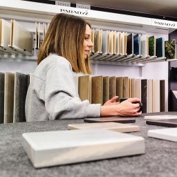 Transmisje na żywo - VisionRed.pl - live streaming online - Kobieta w salonie płytek ceramicznych przegląda próbki płytek, trzymając kubek w dłoni, na tle ekspozycji płytek marki Paradyż.