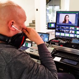 Transmisje na żywo - VisionRed.pl - live streaming online - Operator w słuchawkach i z mikrofonem kontroluje mikser audio-wideo podczas nagrywania wywiadu z kobietą widoczną na ekranach monitorów w tle.