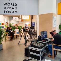 Transmisje na żywo - VisionRed.pl - live streaming online - Nagrywanie panelu dyskusyjnego World Urban Forum: cztery osoby siedzą przy stoliku otoczone roślinami, w tle dekoracja ścienna, a przed nimi kamery na statywach i stanowisko realizatora dźwięku...