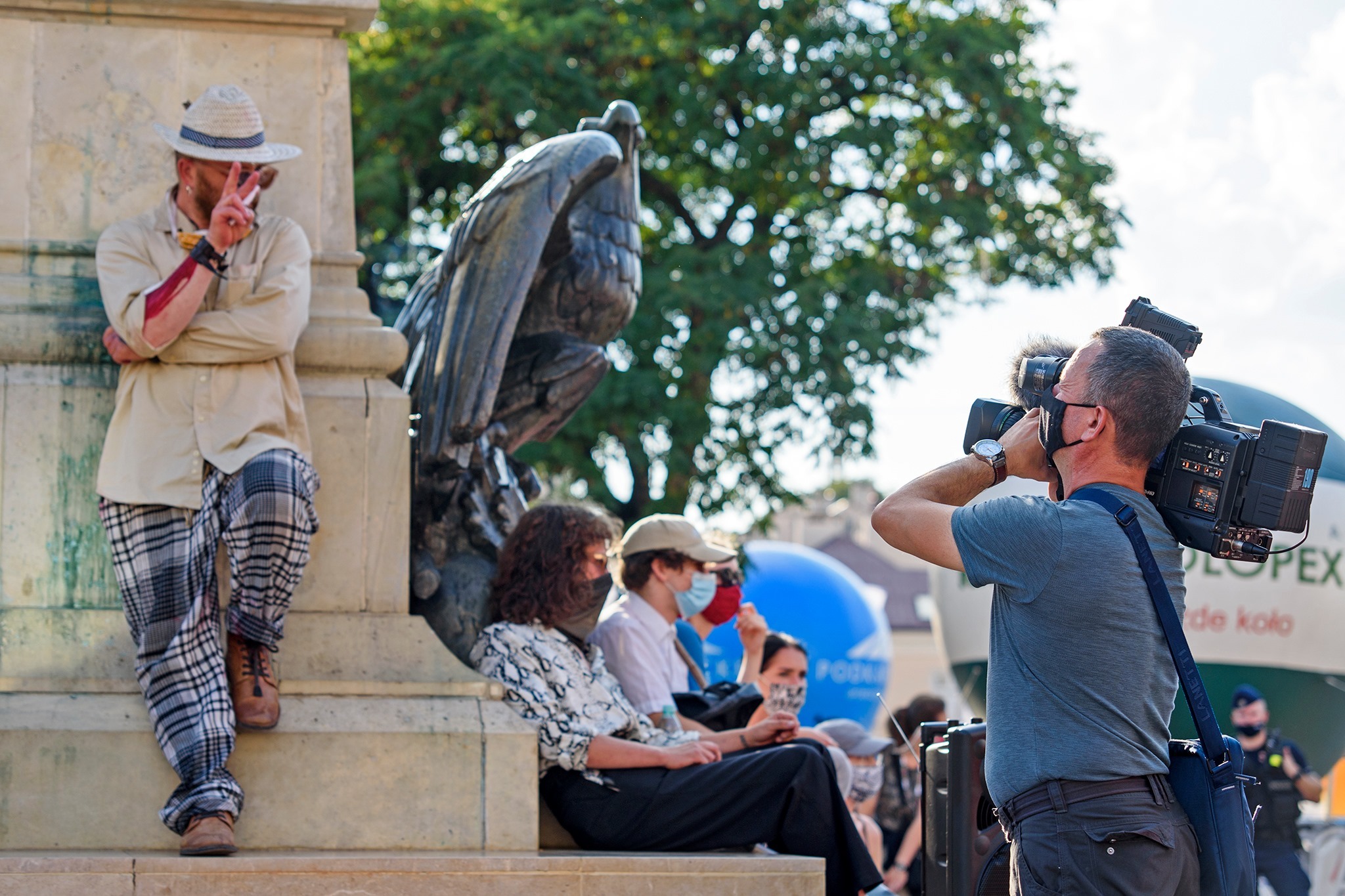 Operator kamery z profesjonalnym sprzętem filmuje osoby siedzące obok pomnika orła. Jedna osoba oparta o pomnik w słomkowym kapeluszu pokazuje znak zwycięstwa.