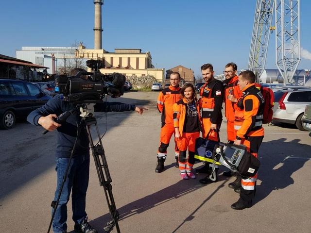 Ekipa filmowa nagrywa materiał z grupą ratowników medycznych w pomarańczowych kombinezonach na tle przemysłowego krajobrazu z kominem i parkingiem.
