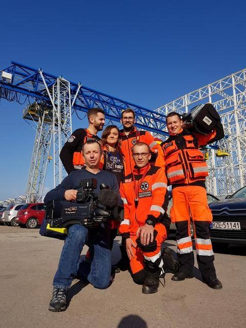 Ekipa filmowa z kamerą i ratownicy medyczni w pomarańczowych kombinezonach pozują na tle dźwigu i parkingu.