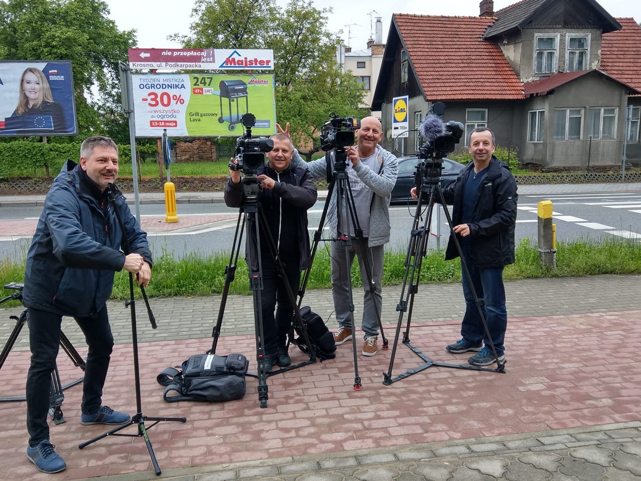 Czterech mężczyzn z profesjonalnymi kamerami na statywach, prawdopodobnie podczas kręcenia materiału w plenerze, billboardy i budynek w tle.