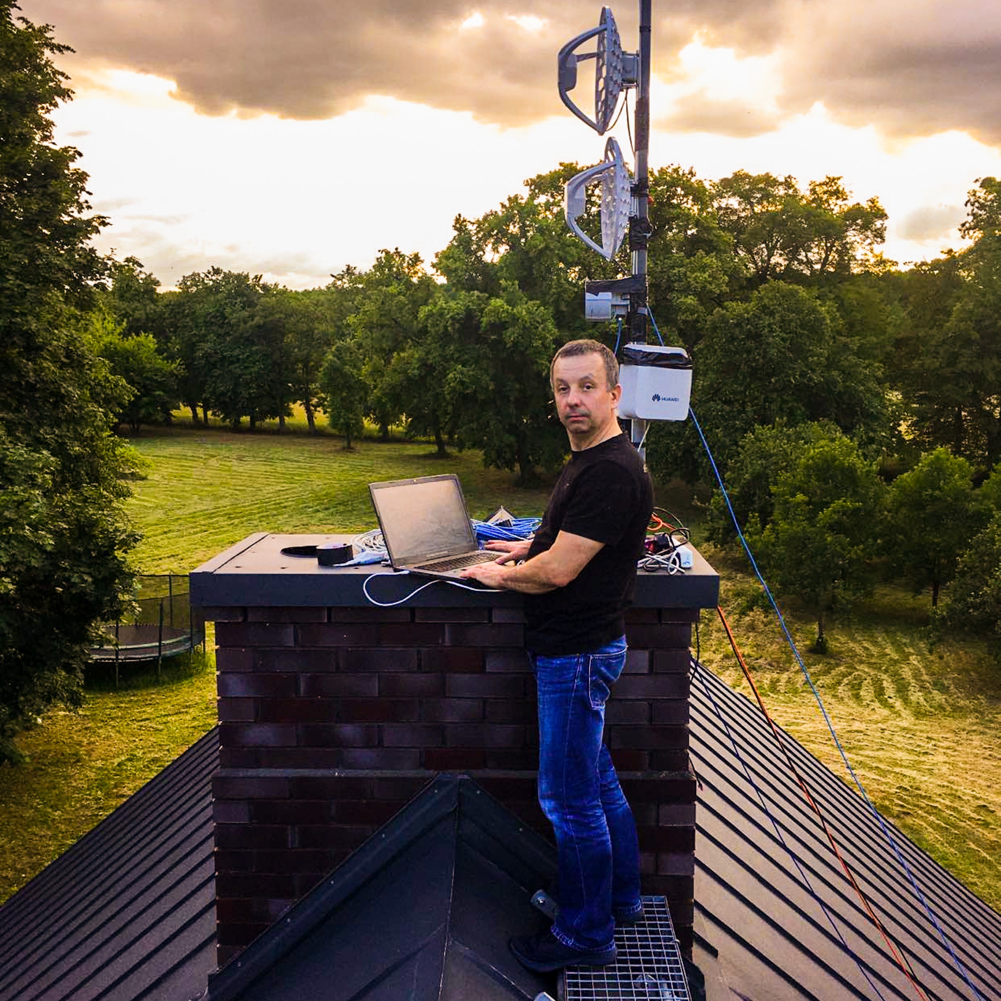 Mężczyzna na dachu komina, konfigurujący antenę internetową za pomocą laptopa; widoczna antena Huawei, kable i trawnik w tle.
