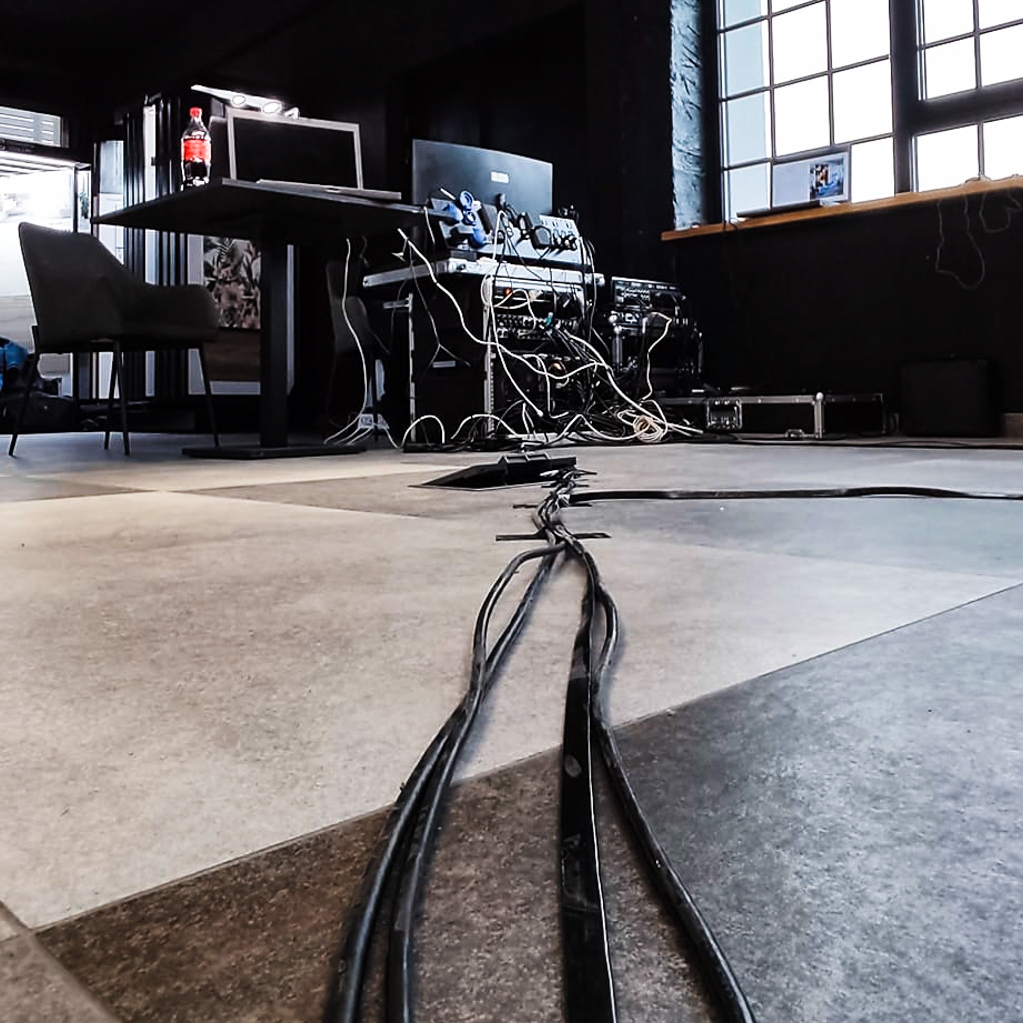 Rozłożone kable zasilające i sygnałowe na podłodze w studio nagraniowym, sprzęt audio-wideo na stoliku w tle, butelka napoju na stole, duże okno z kratami, ciemne ściany.
