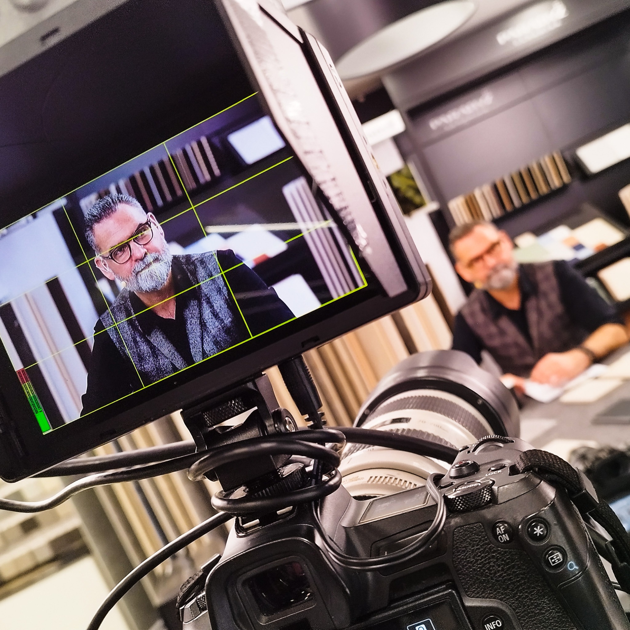 Kamera z podglądem nagrywanego mężczyzny w okularach, ubrany w kamizelkę na tle próbek materiałów wykończeniowych.