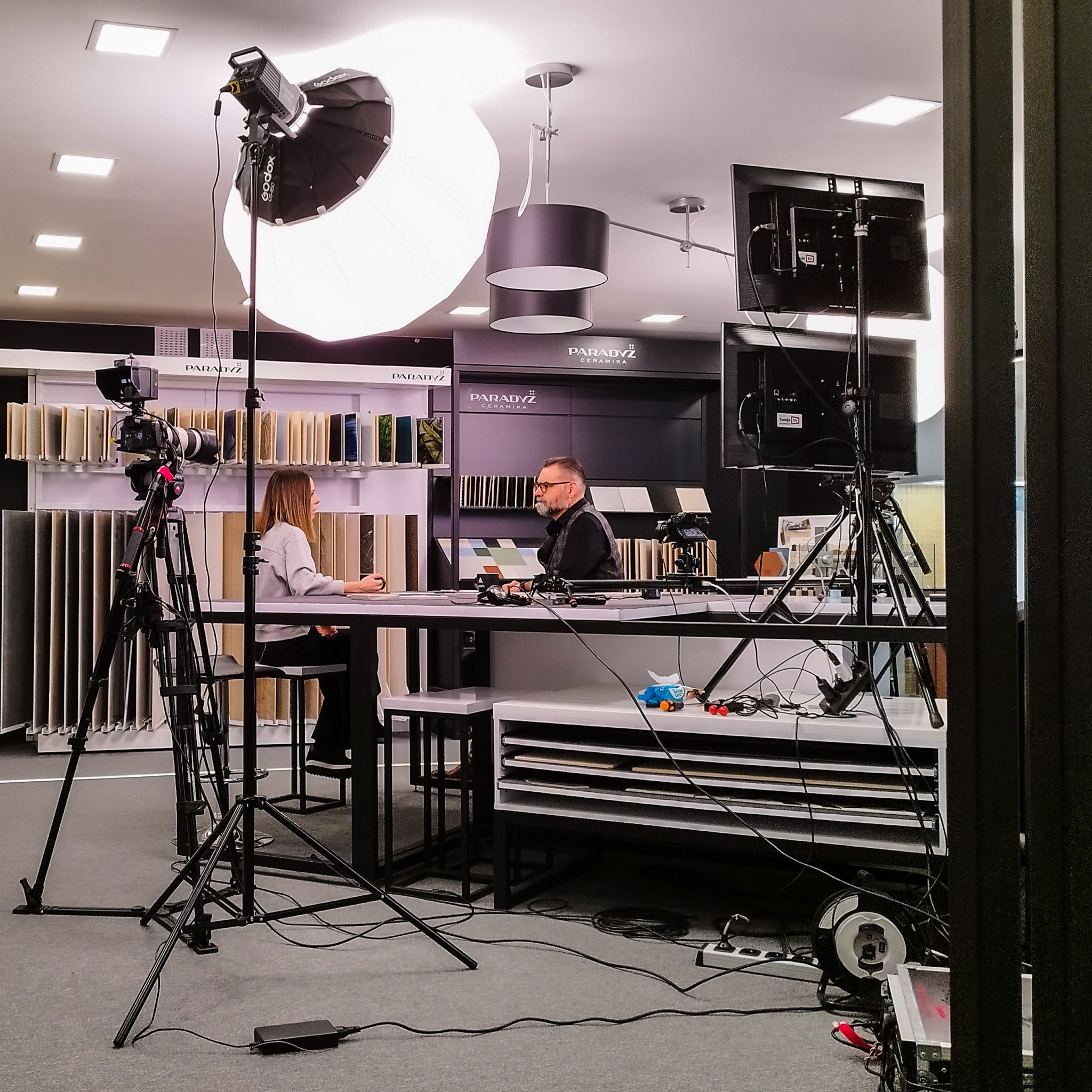 Realizacja nagrania wideo wywiadu w studiu, widoczne oświetlenie studyjne, kamera na statywie, monitory podglądowe, osoba przeprowadzająca wywiad i osoba udzielająca wywiadu na tle ekspozycji...