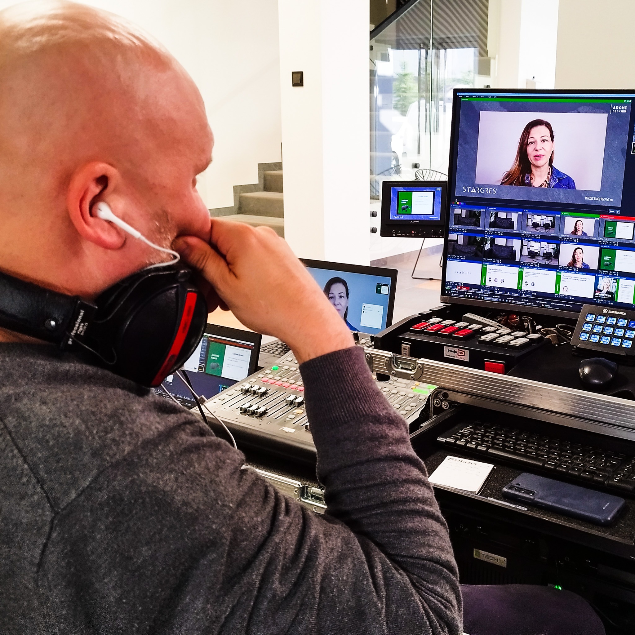 Operator w słuchawkach i z mikrofonem kontroluje mikser audio-wideo podczas nagrywania wywiadu z kobietą widoczną na ekranach monitorów w tle.