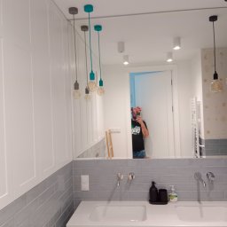 CREATE DK-K - Nowoczesna łazienka z podwójną umywalką, szarą glazurą i białymi szafkami, oświetlona wiszącymi lampami z kolorowymi akcentami, widoczne odbicie osoby robiącej zdjęcie w lustrze.