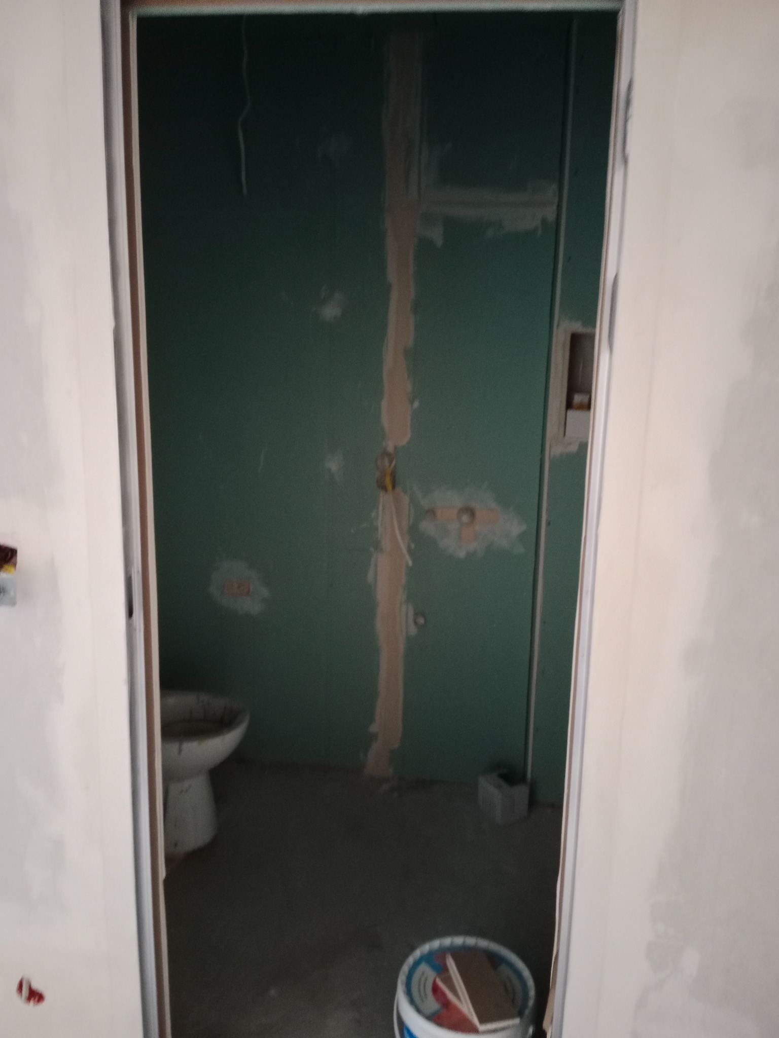 Wnętrze łazienki w trakcie remontu: zielone płyty gipsowo-kartonowe na ścianach, częściowo zaszpachlowane, widoczne instalacje, stara toaleta, wiadro z narzędziami malarskimi w rogu.