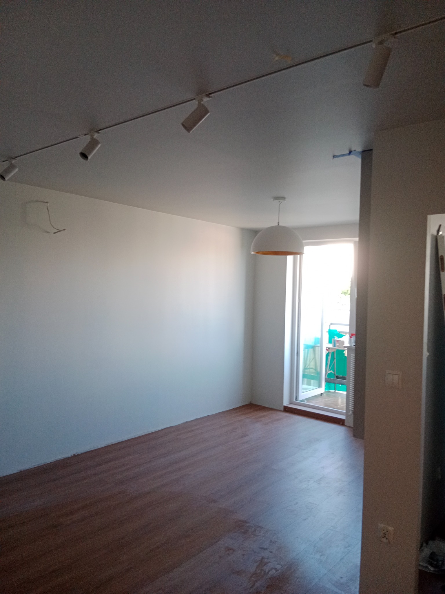 Pusty pokój z drewnianą podłogą, białymi ścianami i reflektorami szynowymi na suficie. Widoczne okno balkonowe z widokiem na balkon.