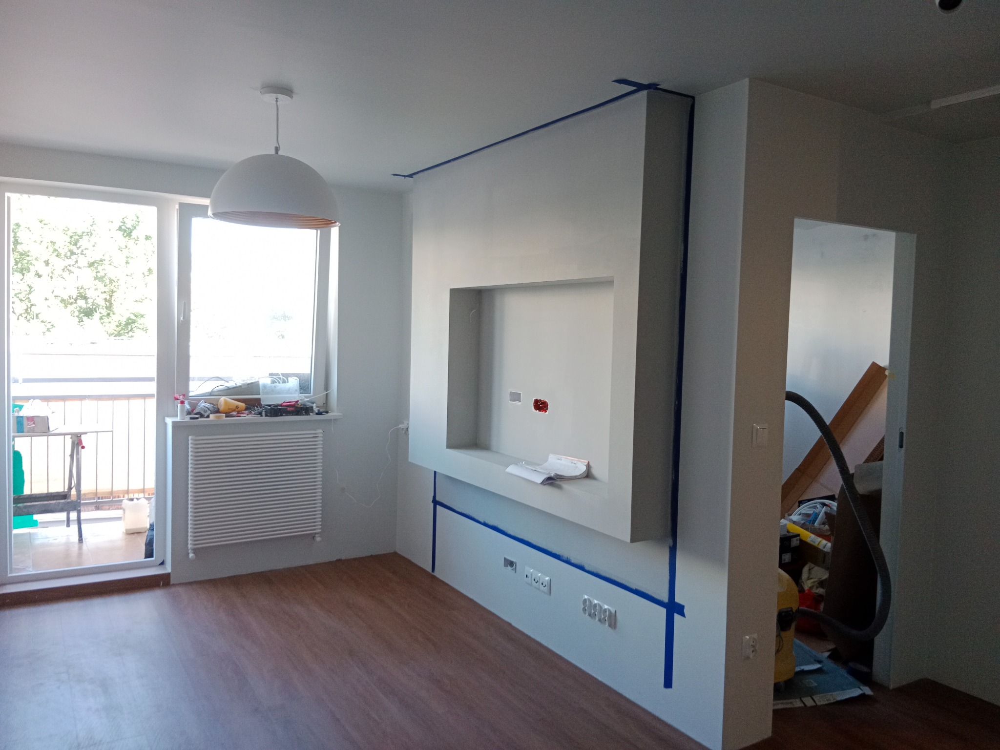 Pokój w trakcie prac wykończeniowych: ściany zabezpieczone taśmą malarską, zabudowa ściany z wnęką na telewizor, widoczne gniazdka elektryczne i instalacja, podłoga z paneli, lampa sufitowa...