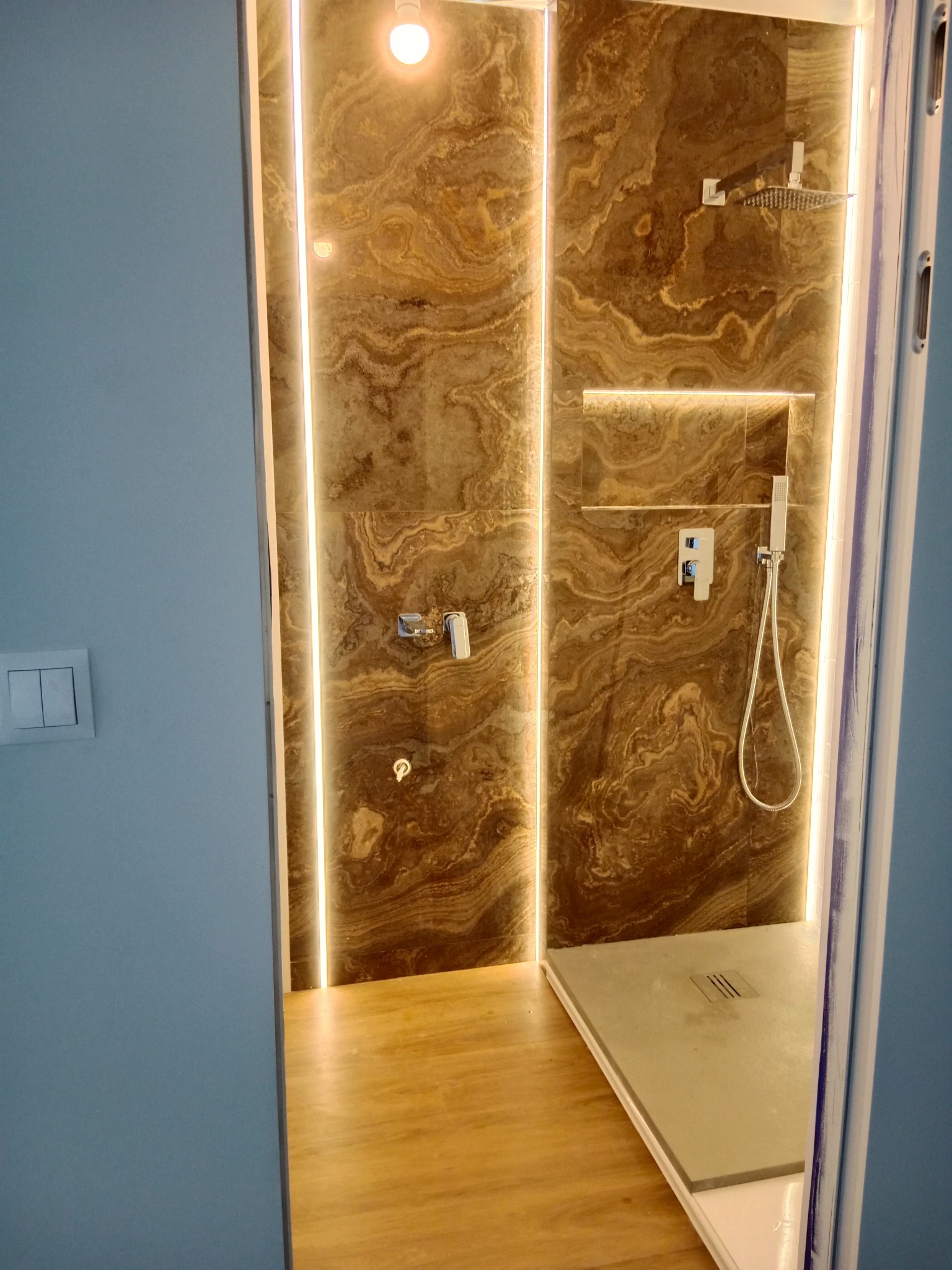 Nowoczesna kabina prysznicowa z brązowymi płytkami imitującymi marmur, podświetlona listwami LED, z drewnianą podłogą i odpływem liniowym w szarej posadzce.