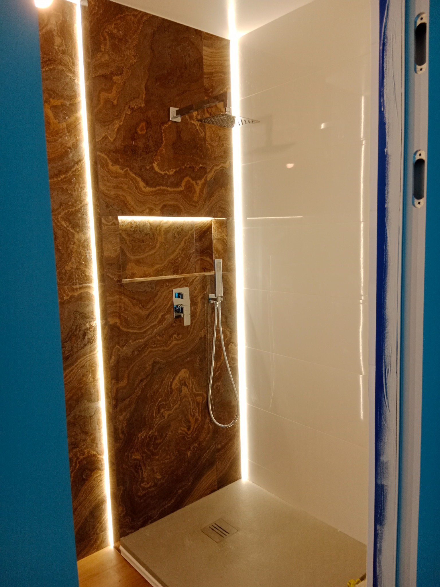 Nowoczesna kabina prysznicowa z brązowymi płytkami imitującymi marmur, podświetleniem LED wzdłuż krawędzi, półką wnękową i białą ścianą boczną wyłożoną płytkami.