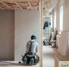 Pracownik w kasku i białej koszulce, z narzędziami przy pasie, siedzi na podłodze w surowym wnętrzu i przygotowuje ścianę do dalszych prac wykończeniowych. Widoczne płyty gipsowo-kartonowe...