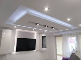 Biały sufit podwieszany z oświetleniem LED i halogenami, w tle białe meble i okno z zasłonami.