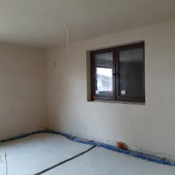 Pokój po nałożeniu tynku maszynowego, widoczne ściany z nałożonym tynkiem, okno z roletami, wystające kable oraz rury ogrzewania podłogowego.