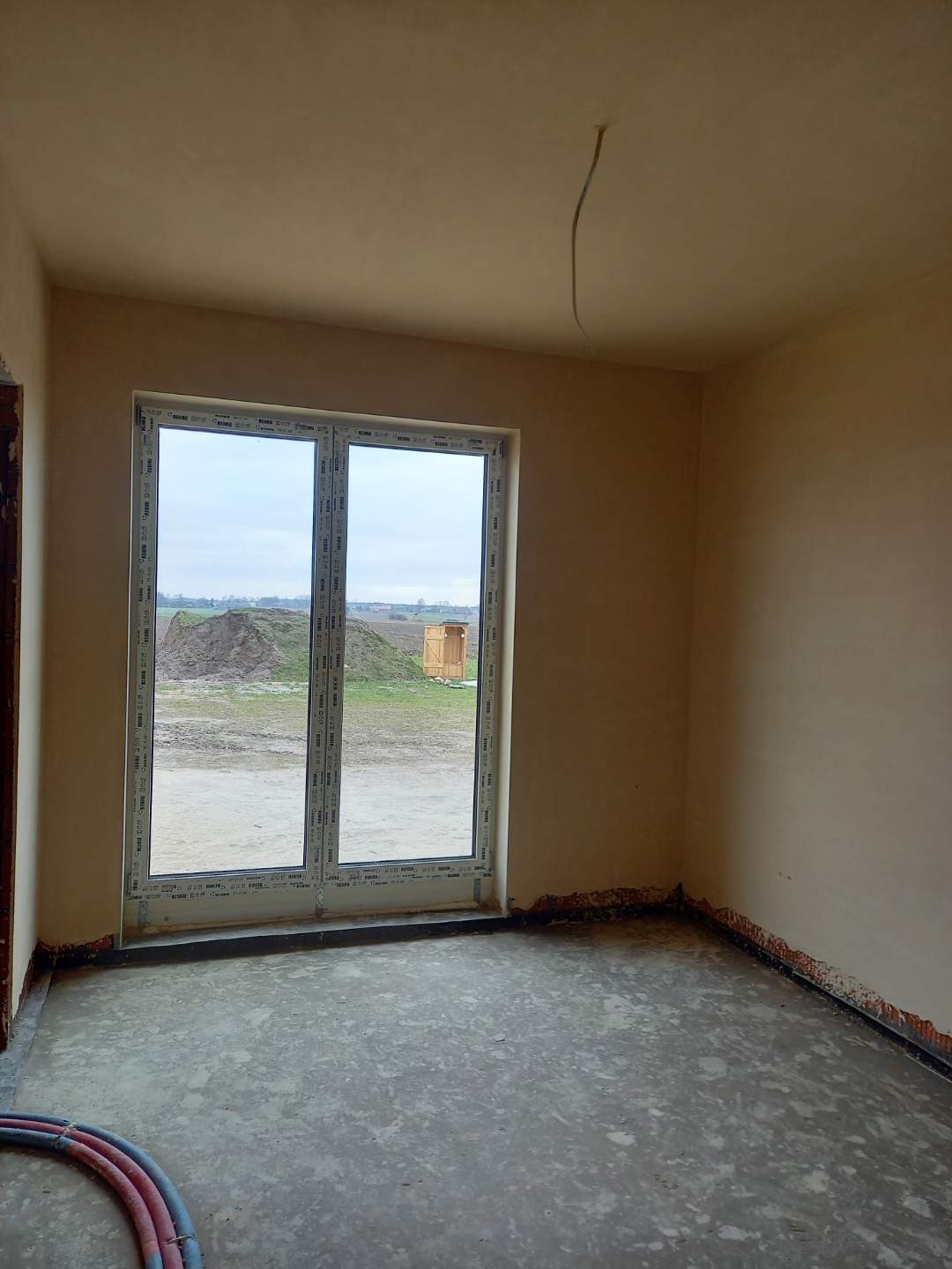 Wnętrze surowego pomieszczenia z nałożonymi tynkami, widoczne ślady po pracach wykończeniowych na podłodze i przy oknach, wystający kabel z sufitu, oraz dwuskrzydłowe okno z widokiem na pole i mały...